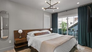 Una habitación de hotel moderna con una cama grande, una mesita de noche con lámpara, un espejo y vistas al balcón exterior.