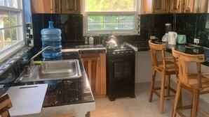 A kitchen with wooden cabinets, a black countertop, a stove, and a window with a view of greenery. - Bright Country Apartment at Robin Hood guesthouse (Port Antonio)