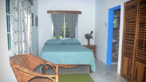 A bedroom with a bed, a wicker chair, a nightstand, and a wooden wardrobe. - Bright Country Apartment at Robin Hood guesthouse (Port Antonio)