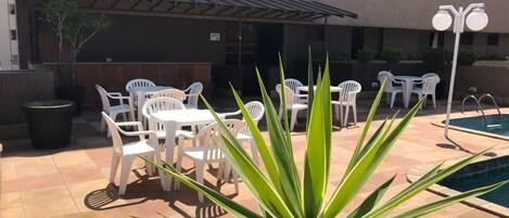 An outdoor patio area with white plastic chairs and tables, a pool, and a large plant in the foreground.