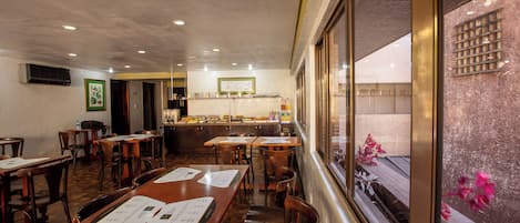 Un restaurante con mesas y sillas de madera, una zona de cocina y vistas a través de una ventana.