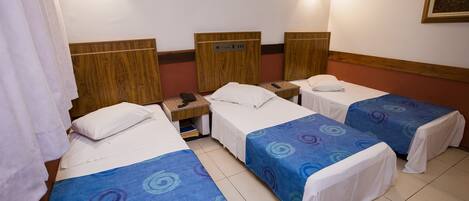 Habitación de hotel con tres camas individuales, cada una con una colcha azul y blanca. Se aprecia un cabecero de madera a la izquierda, una mesita de noche con un teléfono y una lámpara fijada a la pared.