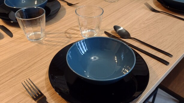 A dining table set with blue plates, black bowls, and clear glasses, accompanied by cutlery and a spoon.