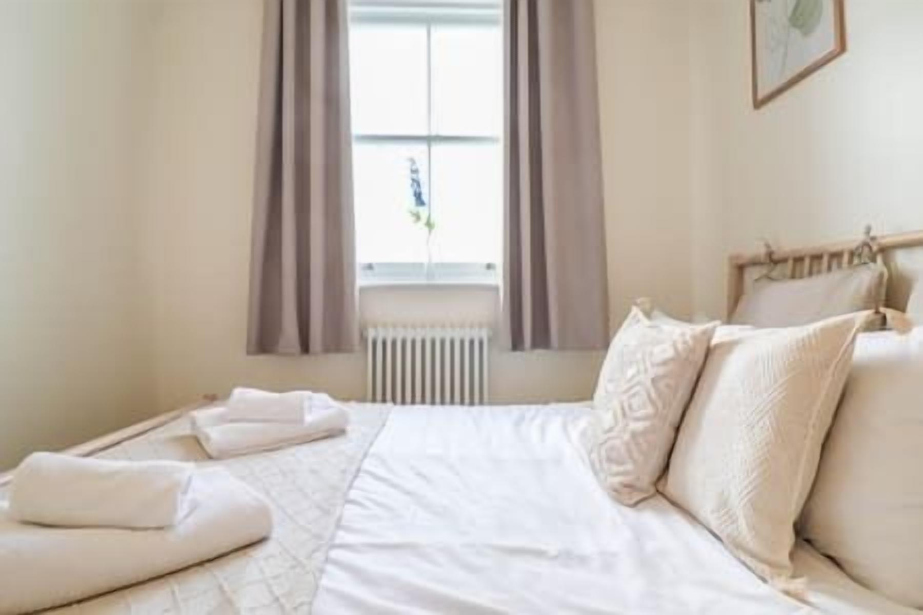 A neatly made bed with white linens and pillows, a window with curtains, and a radiator below.