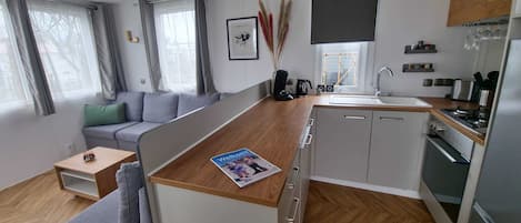 Un espace de vie moderne et compact, doté d’une kitchenette, d’un canapé gris, d’un plancher en bois et offrant une vue sur l’extérieur.