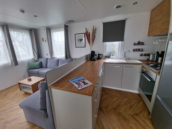 A modern, compact living space with a kitchenette, grey sofa, wooden flooring, and a view of the outside. - Modernes, Junges Chalet mit Terrasse. Haustierfreundlich! (Oostkapelle)