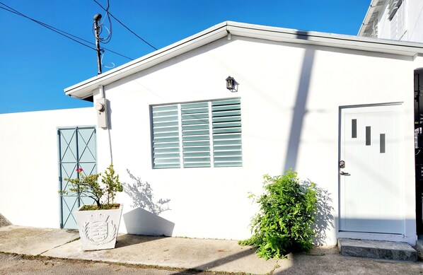 Un bâtiment blanc avec une seule fenêtre, une porte et un petit jardin agrémenté d’une plante en pot.