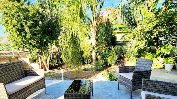 Un espace extérieur aménagé en terrasse, avec des meubles en osier et un sol en pierre, entouré d’une végétation luxuriante et d’arbres.