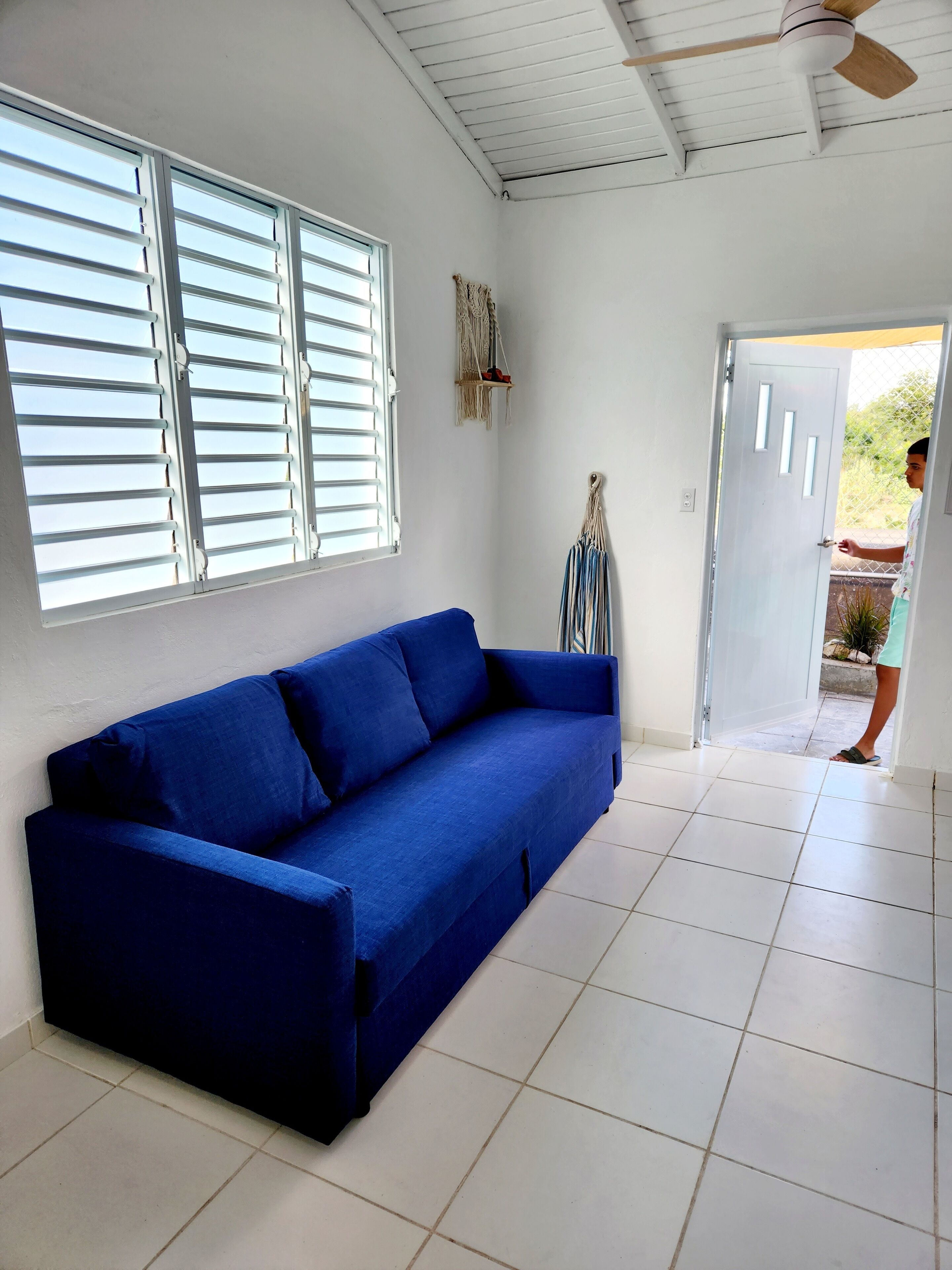 Una habitación con un sofá azul, suelo de baldosas blancas, un ventilador de techo y una puerta que da al exterior.