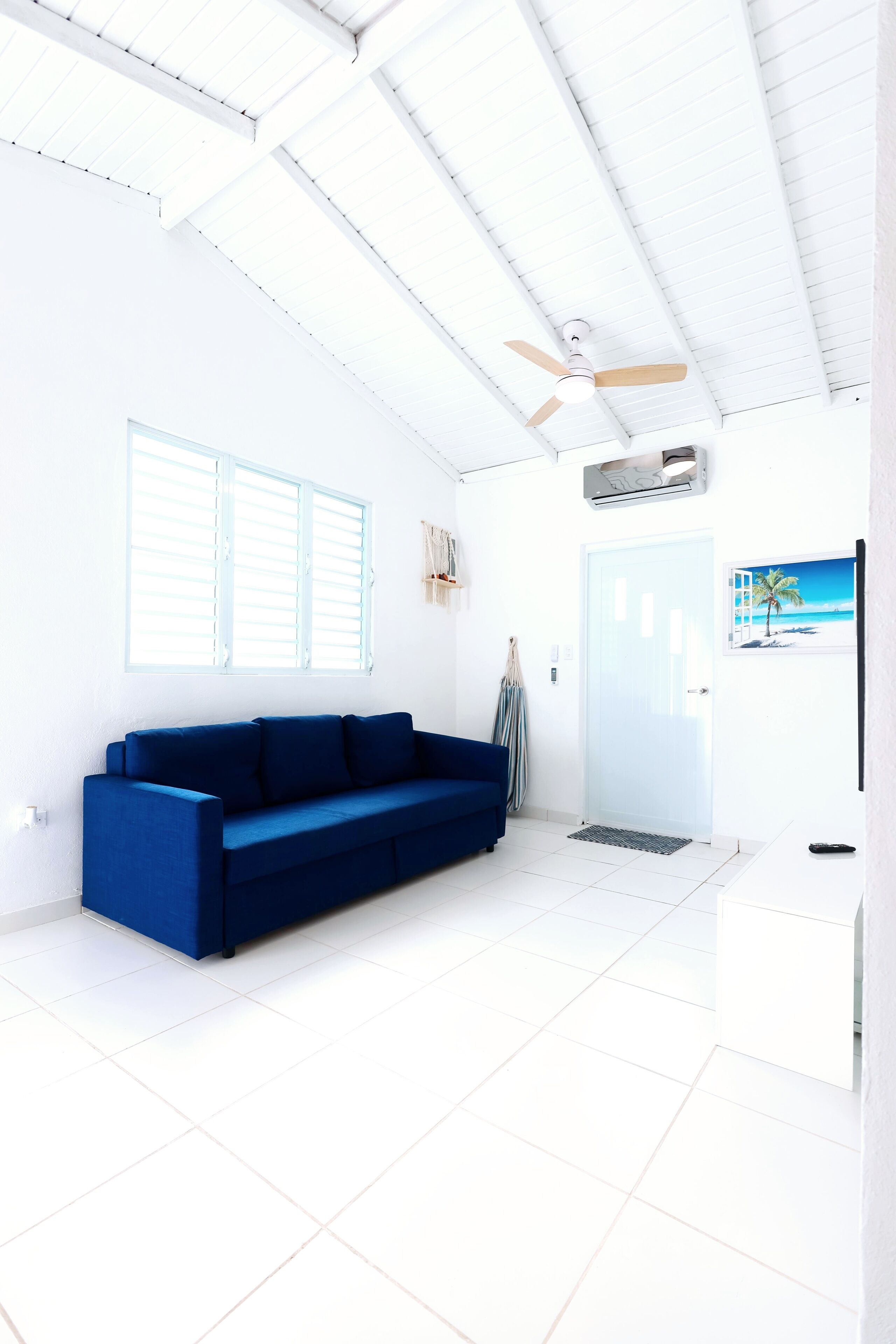 Ein helles Zimmer mit weißer Decke, einem blauen Sofa, einer Tür und einem an der Wand befestigten Fernseher.