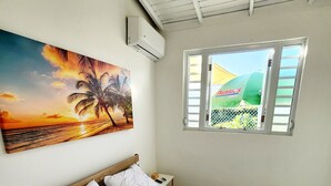 Une chambre à coucher avec un lit, un tableau représentant un coucher de soleil, une fenêtre et un appareil de climatisation.