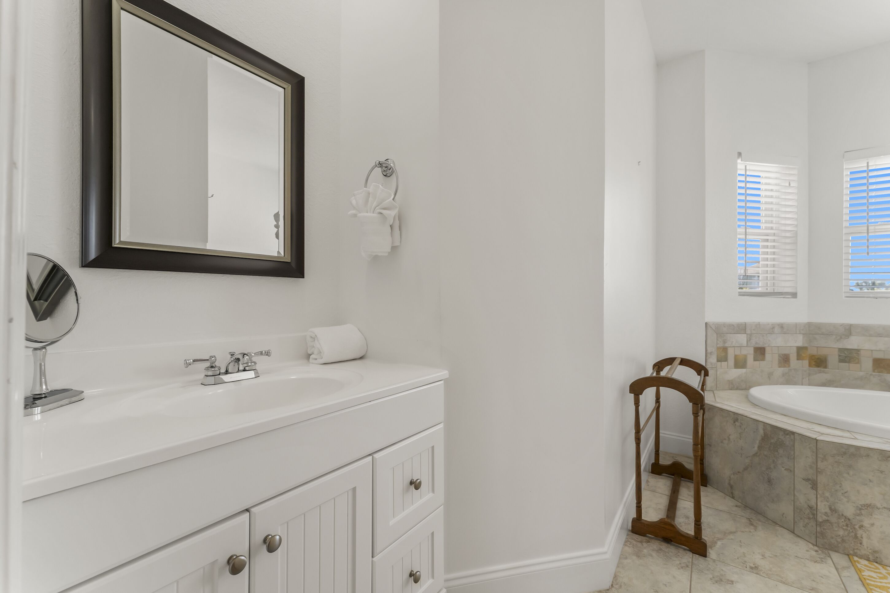 Une salle de bain avec un meuble-lavabo blanc, un grand miroir, un porte-serviettes et une baignoire dont les parois sont recouvertes de carreaux.