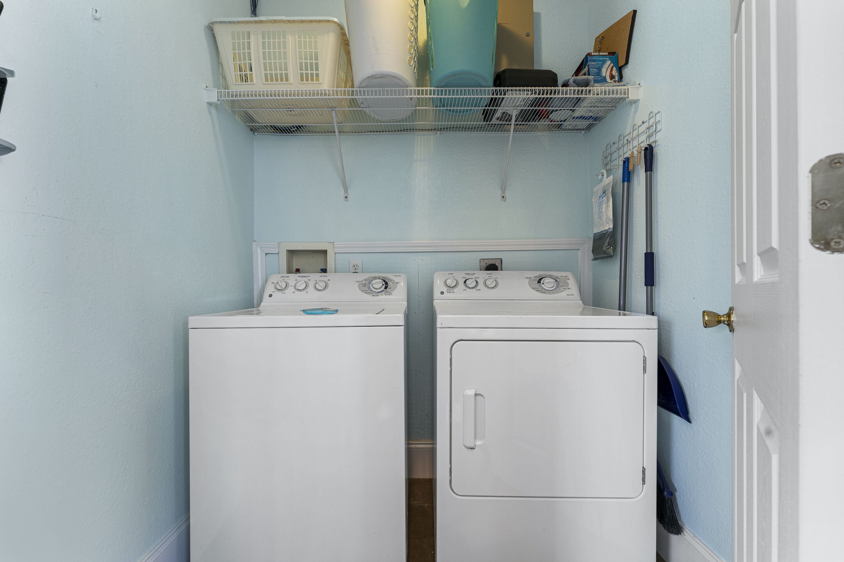 A laundry room with two white washing machines, a shelf above holding laundry baskets and cleaning supplies, and a door to the right.