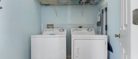 A laundry room with two white washing machines, a shelf above holding laundry baskets and cleaning supplies, and a door to the right.