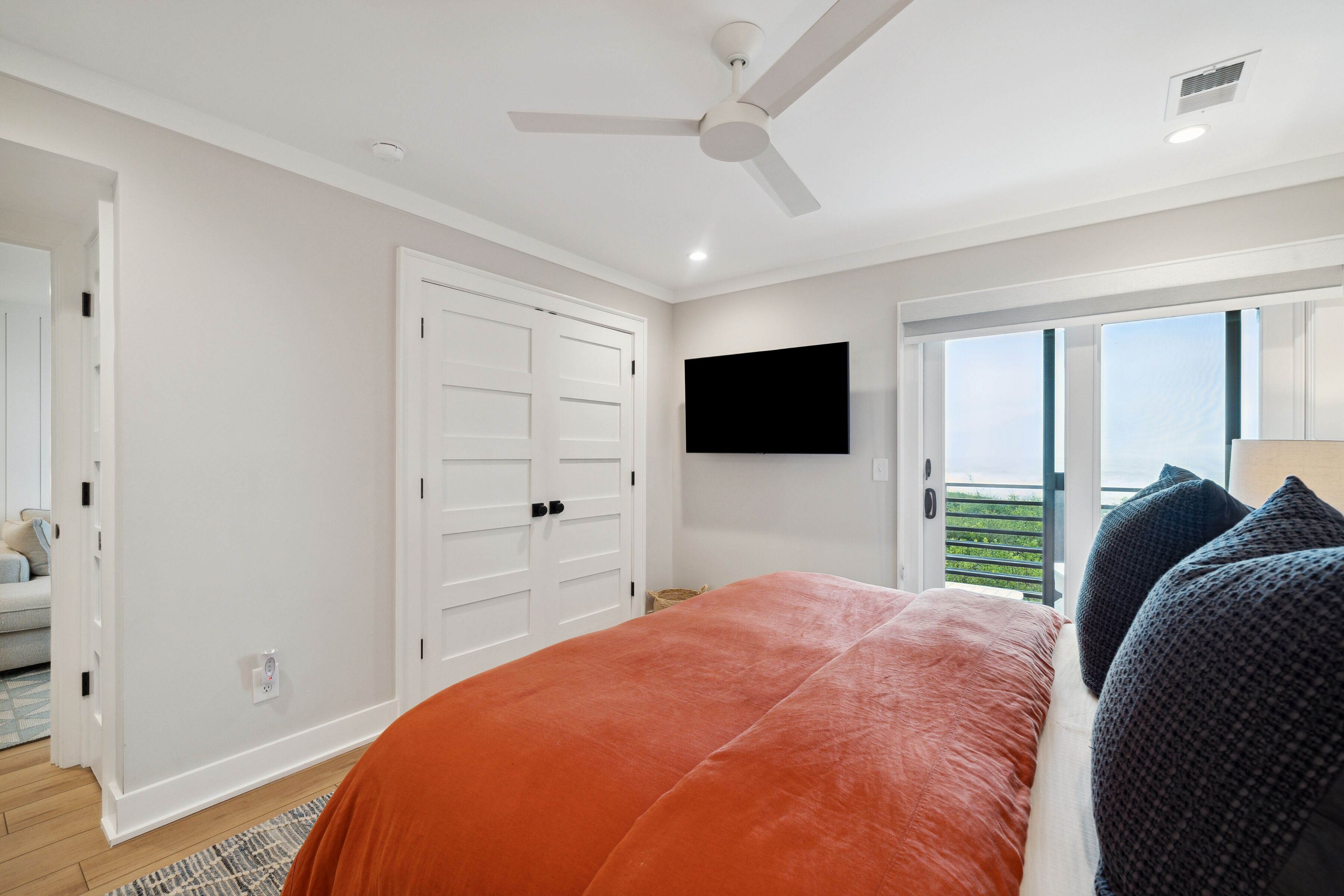 A bedroom with a bed, a ceiling fan, a TV mounted on the wall, and a sliding glass door leading to a balcony.