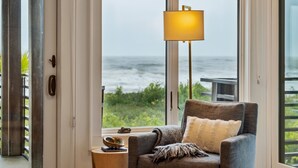 Une pièce avec vue sur l’océan, un fauteuil gris avec un plaid, une lampe sur pied et une petite table sur laquelle se trouvent un bol et un livre.