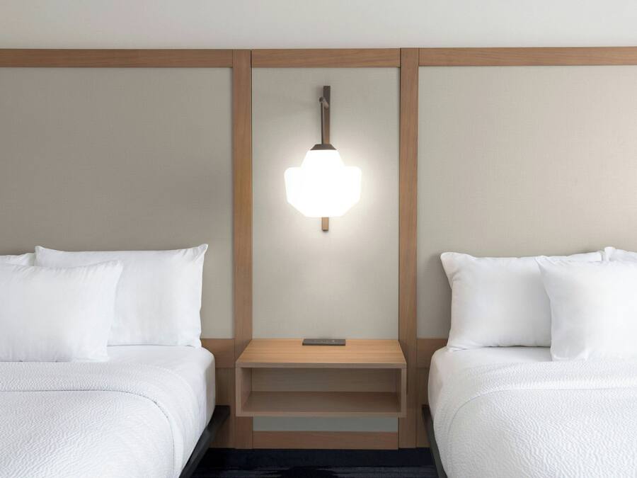 Hotellihuone, jossa on kaksi sänkyä, yöpöytä, seinään kiinnitetty valaisin ja puupanelointi.