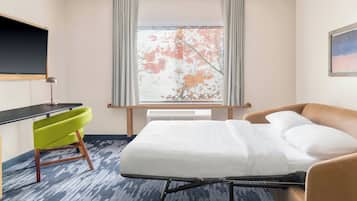 Habitación de hotel con cama, escritorio, una silla verde, ventana con hojas otoñales y un cuadro en la pared.