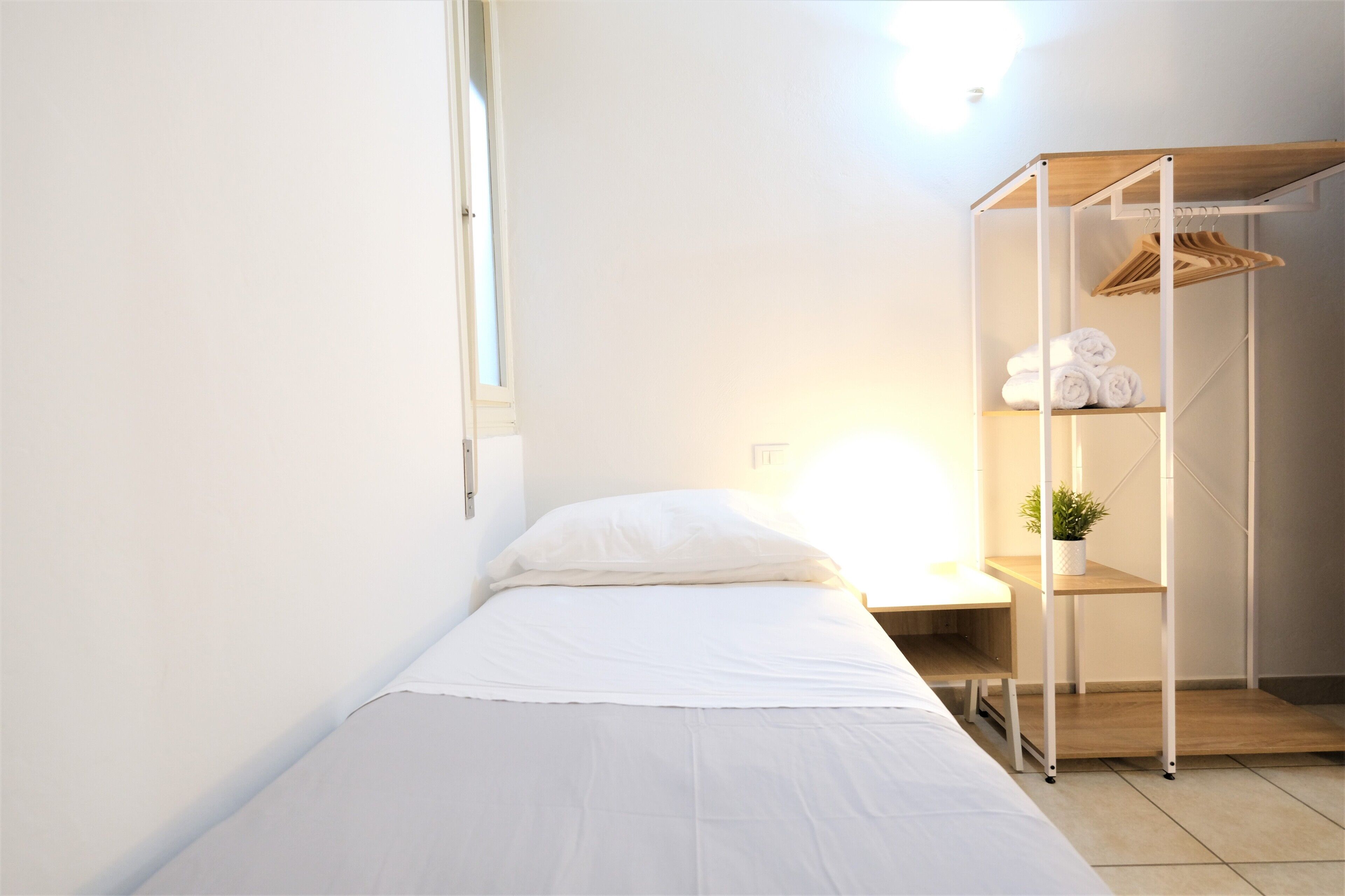 A minimalist bedroom with a single bed, a wooden wardrobe, and a small desk with a plant.