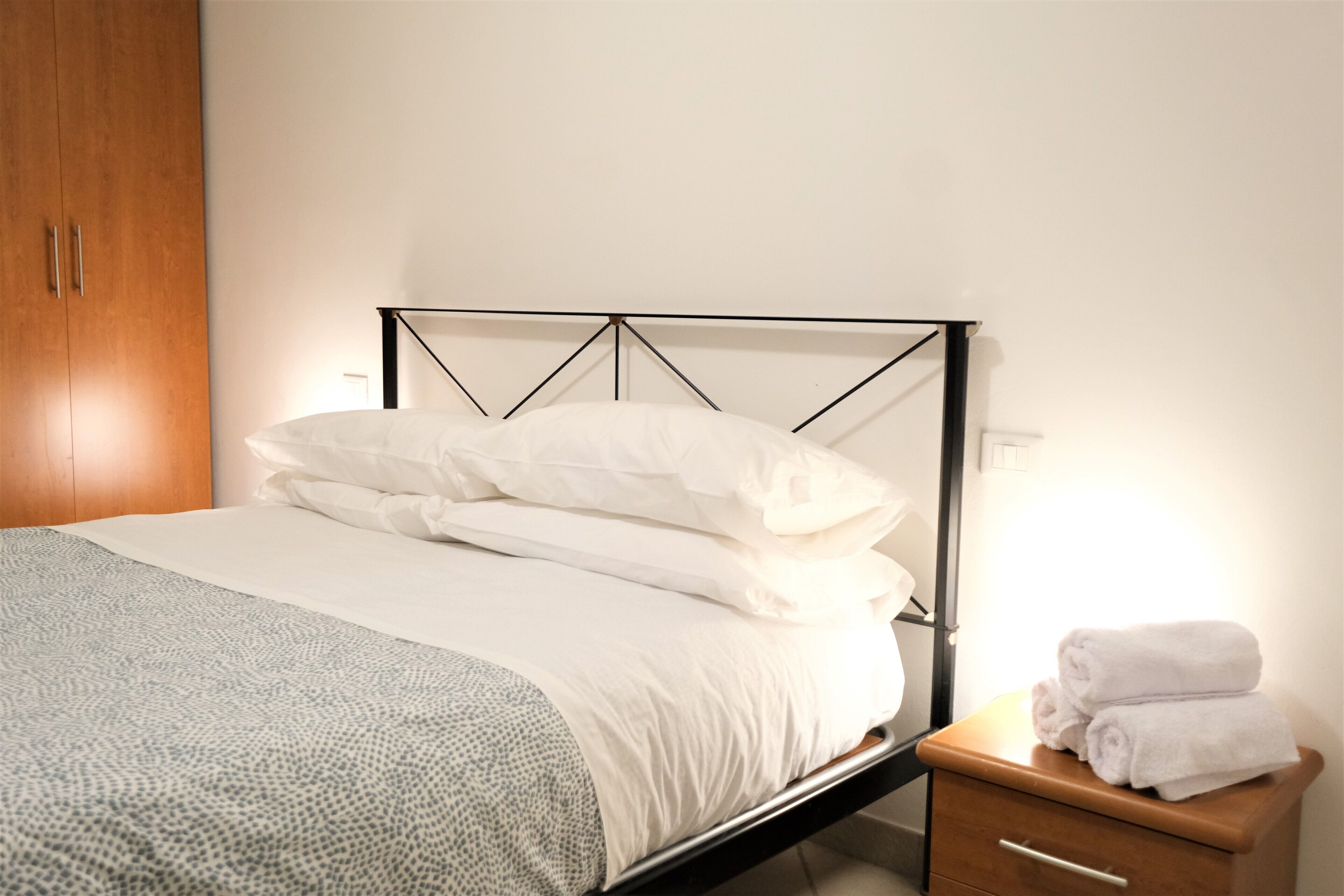 Una cama bien hecha con sábanas blancas, una mesita de noche de madera con un cajón y un armario de madera.