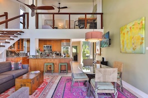 A spacious living area with a high ceiling, a kitchen with wooden cabinets, a dining area with a table and chairs, and a staircase leading to an upper level. - Snorkel with sea turtles! Ocean Front & Pool! (Lahaina)