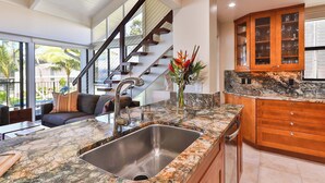 A modern kitchen with a granite countertop, stainless steel sink, wooden cabinets, and a staircase leading to an upper level. - Snorkel with sea turtles! Ocean Front & Pool! (Lahaina)