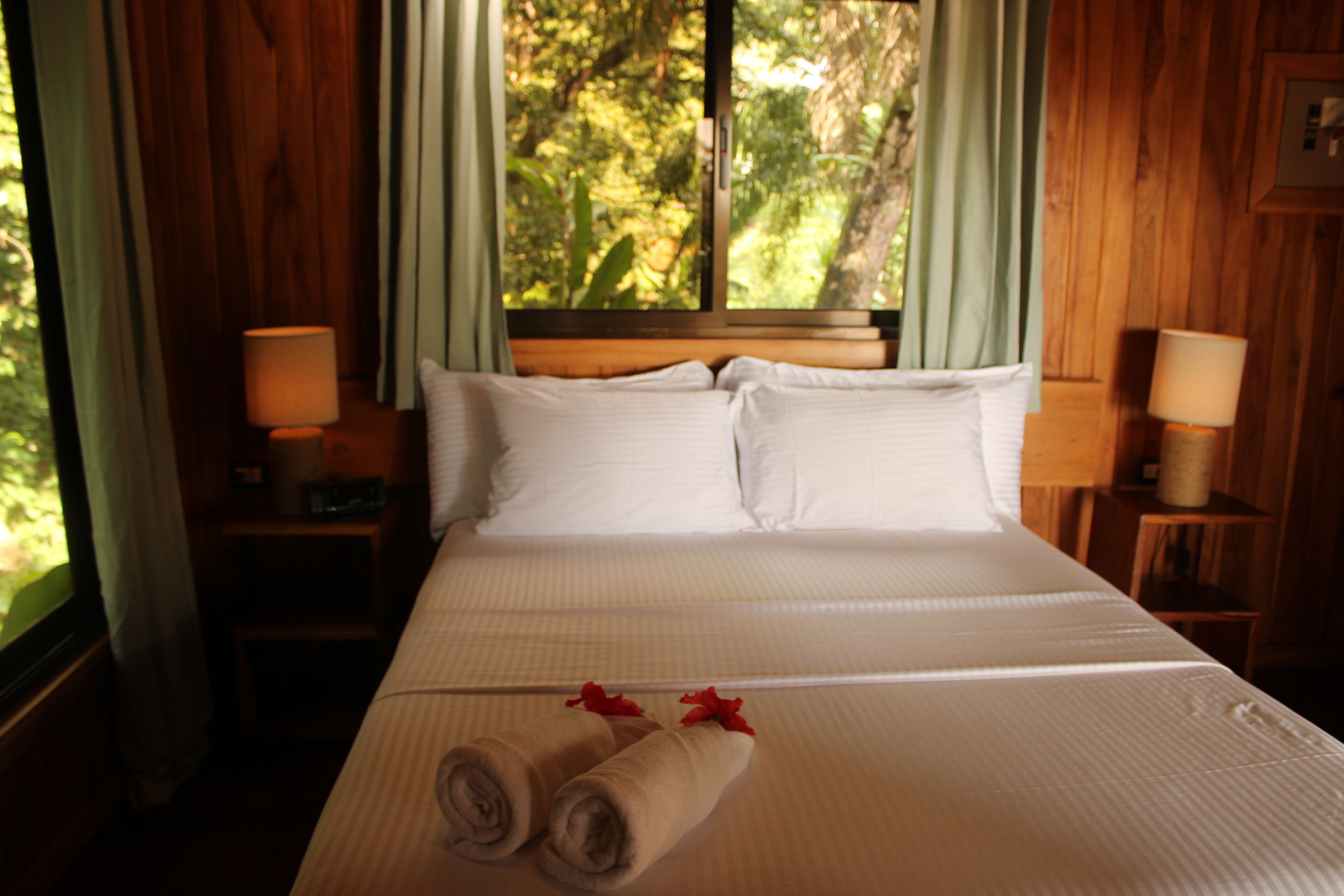 A hotel room with a large bed, wooden headboard, two bedside tables with lamps, and a window with curtains.