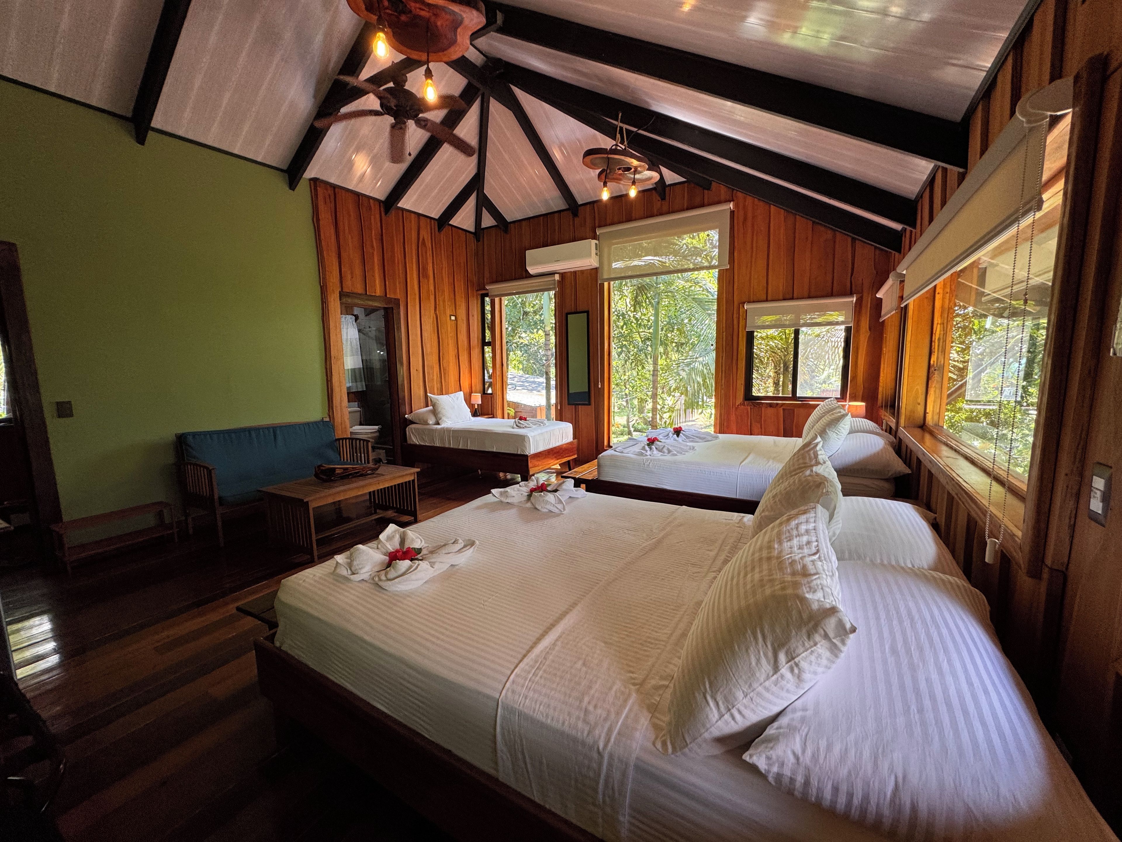 A room with two beds, a wooden ceiling, and a window with a view of greenery.