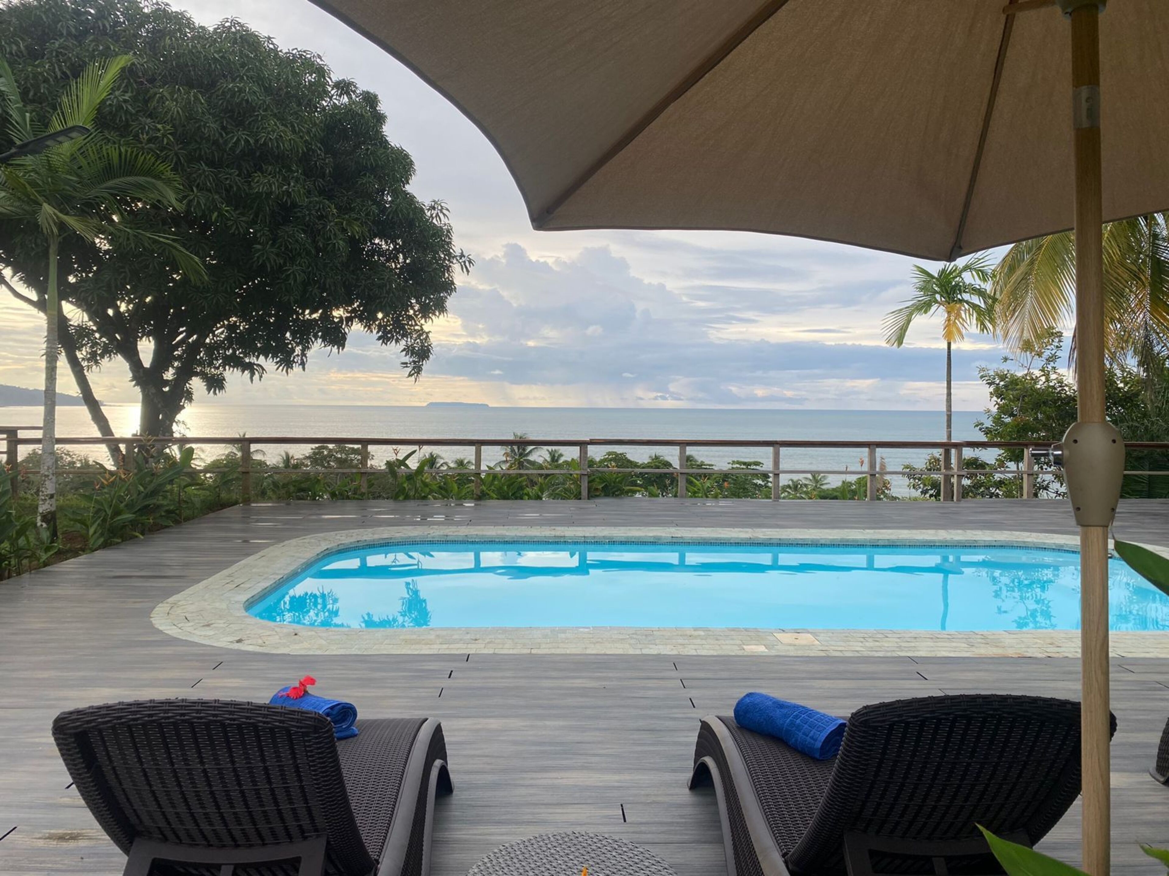 Zona junto a la piscina con vista al océano, muelle de madera y sillones de descanso.