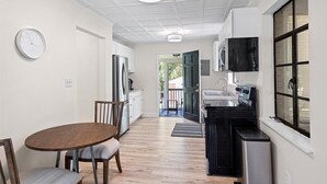 A compact kitchen with a dining area, featuring a wooden table and chairs, a clock on the wall, and a view into the backyard through an open door. - Noda Home w Yard Perfect For Digital Nomads 250 Mbps Fiber! (Charlotte)