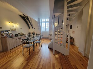 Un coin repas avec une table et des chaises en bois, un escalier blanc et une fenêtre laissant entrer la lumière naturelle.