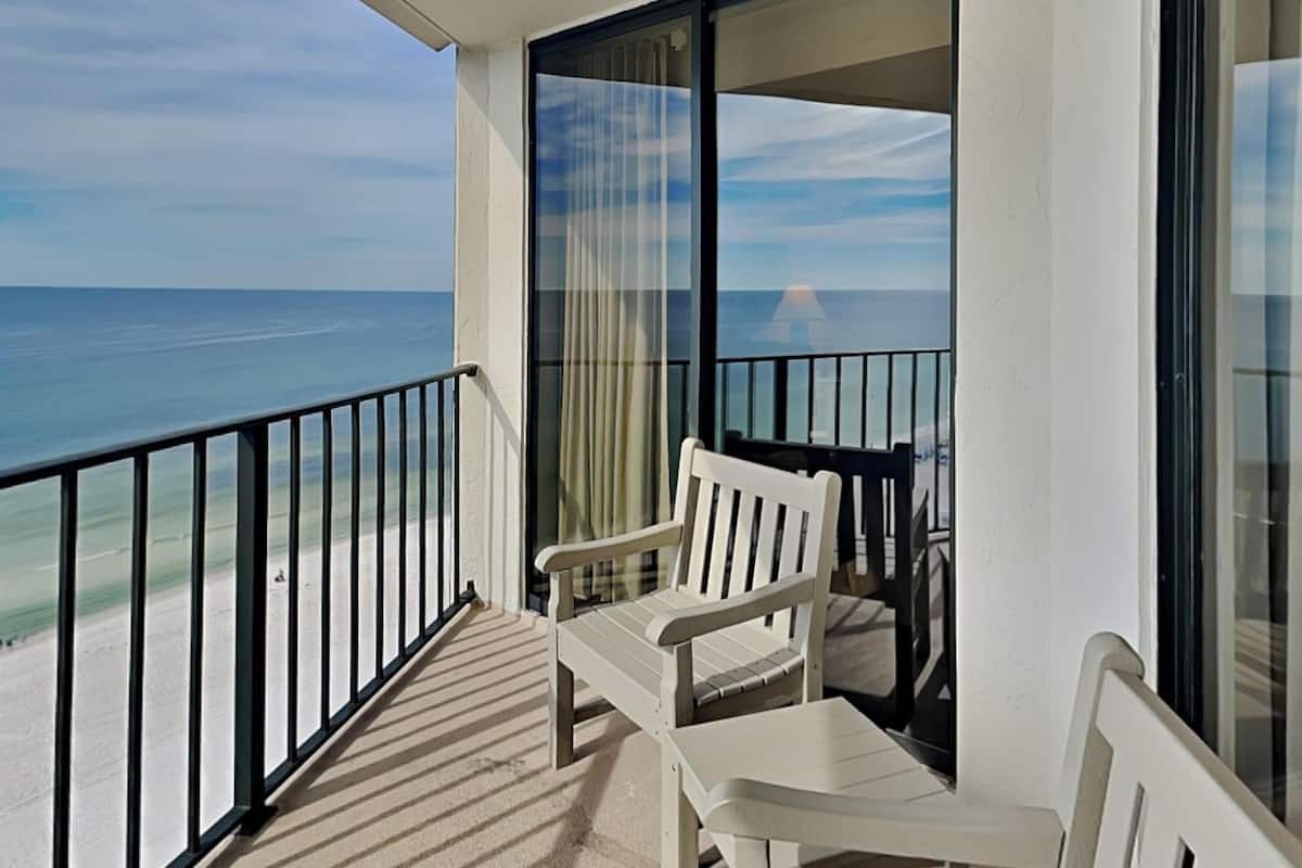 Un balcon avec une chaise et une table blanches, donnant sur une plage et l’océan.