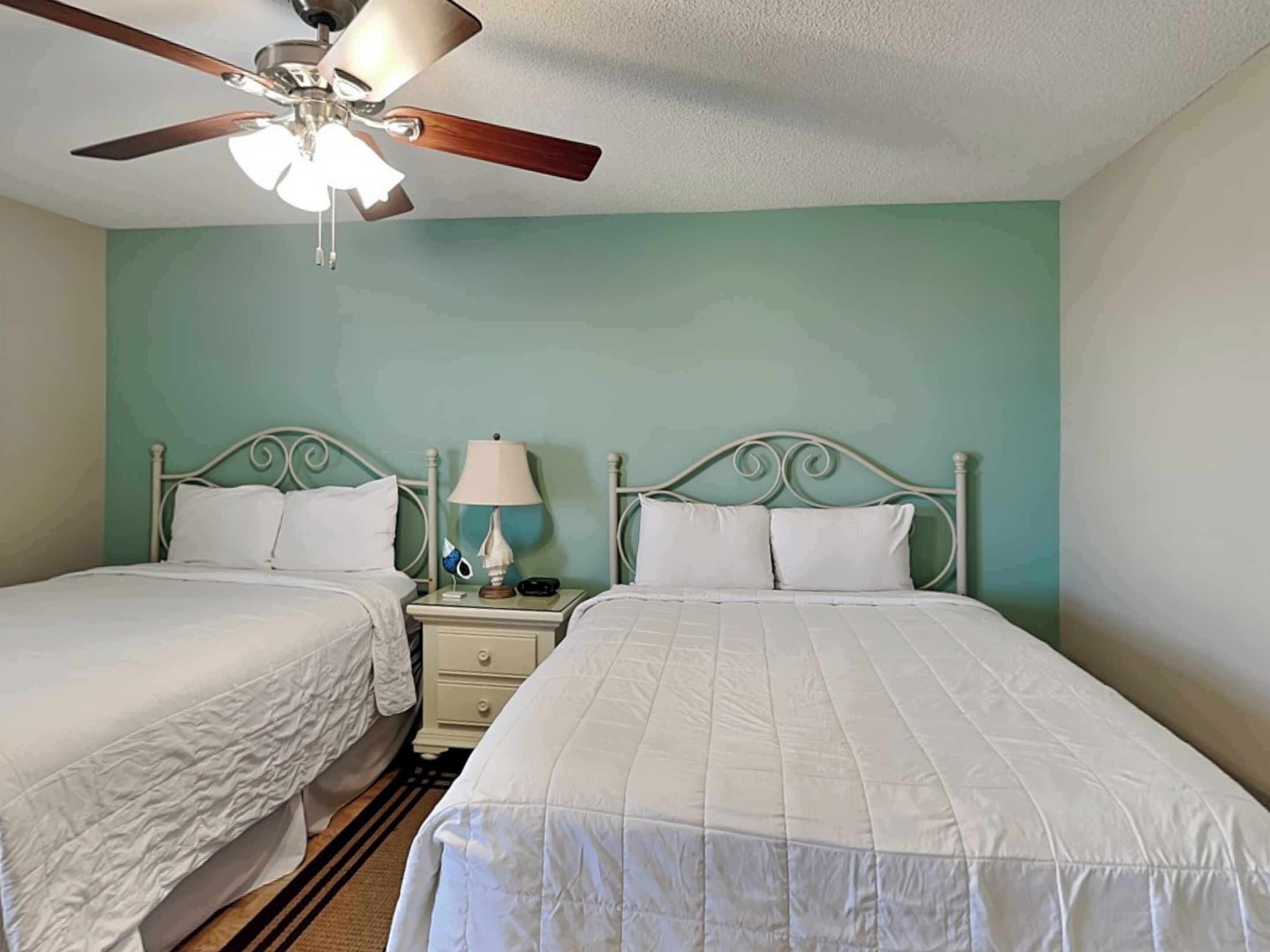 A twin room with two beds, a ceiling fan, a nightstand with a lamp, and a wall clock.