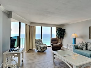 A living room with a large window, a television, a sofa, a coffee table, and a plant. - Edgewater Tower II by Resort Collection (Panama City Beach)