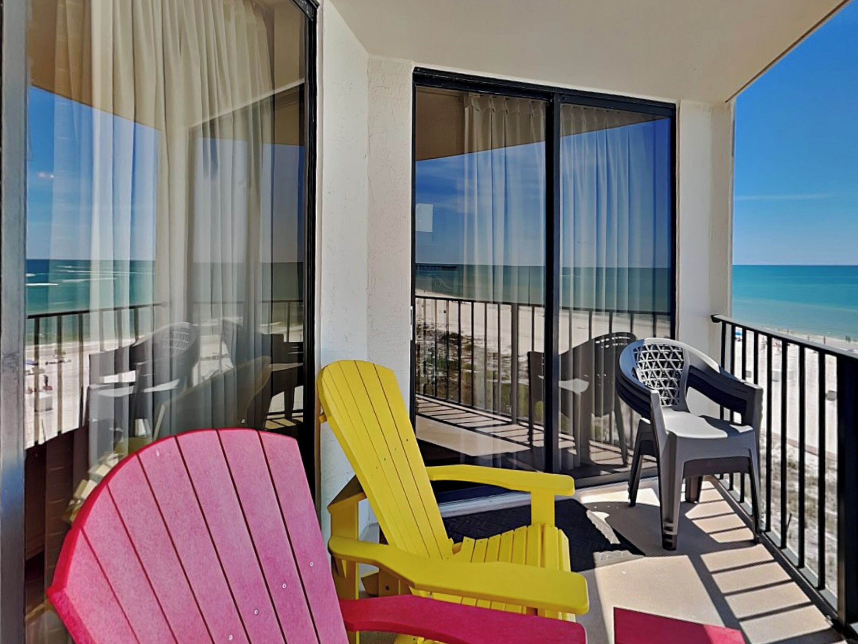 A balcony with a view of the beach, featuring two Adirondack chairs and a small table.