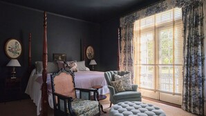 Une chambre à coucher avec un lit à baldaquin, un fauteuil, un repose-pieds et une grande fenêtre avec des rideaux.