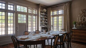 Une salle à manger avec une grande table en bois, dressée pour six personnes, entourée de chaises. De grandes fenêtres sont ornées de rideaux, une bibliothèque expose de la porcelaine, et un lustre illumine la pièce.