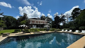 Une piscine entourée de chaises longues et de parasols, elle-même bordée d’un jardin bien entretenu et d’un grand bâtiment en briques doté d’un balcon.
