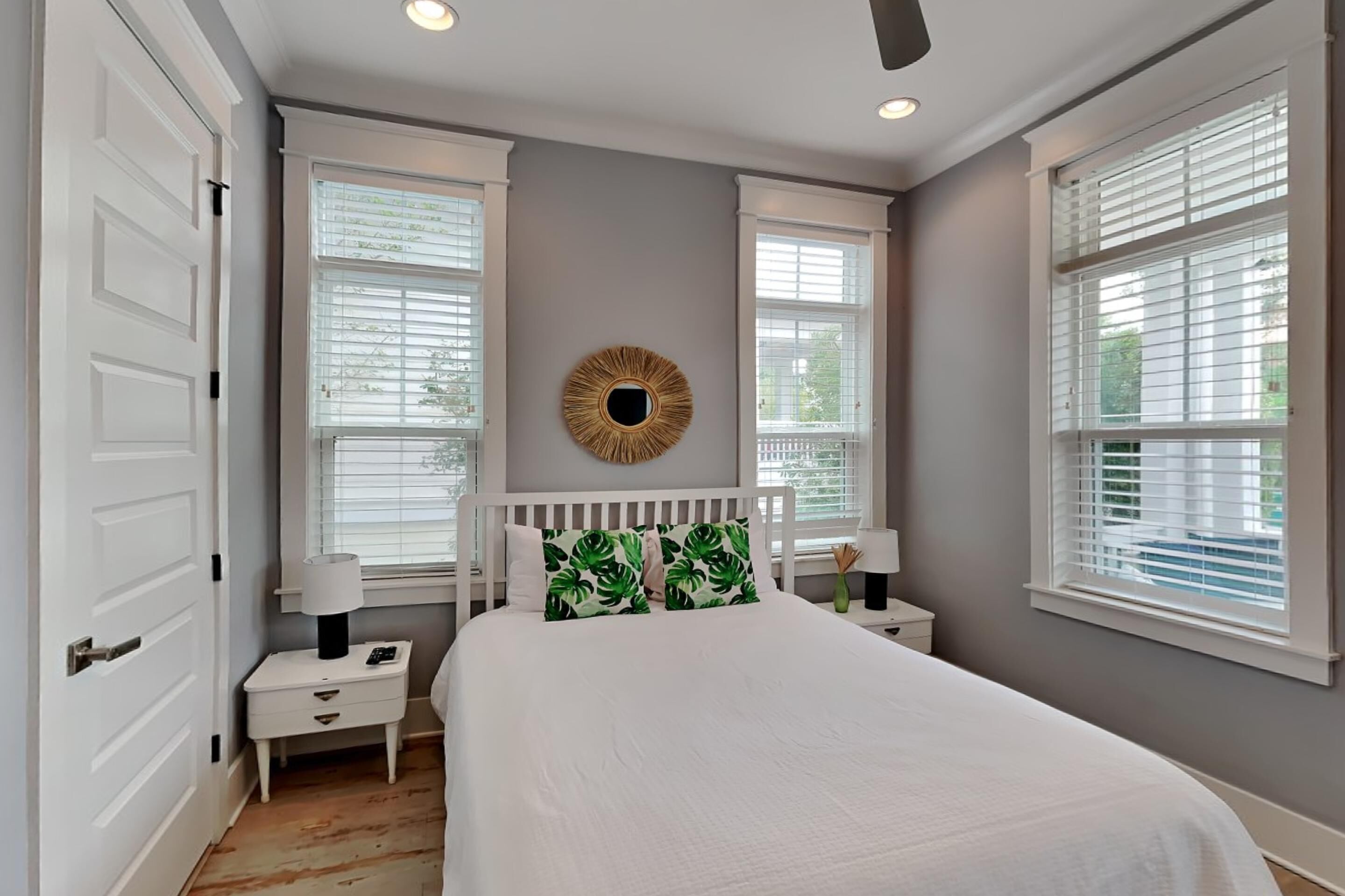 A bedroom with a large bed, two bedside tables, a nightstand, a round mirror, and two windows with blinds.