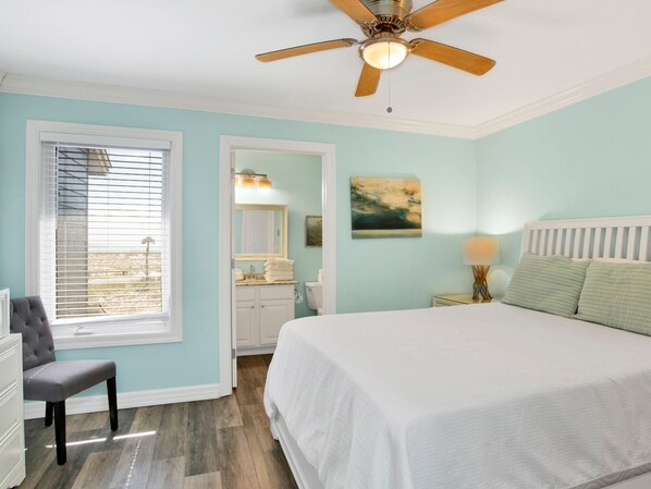 A bedroom with a bed, a ceiling fan, a window with blinds, a chair, a nightstand with a lamp, and a painting on the wall.