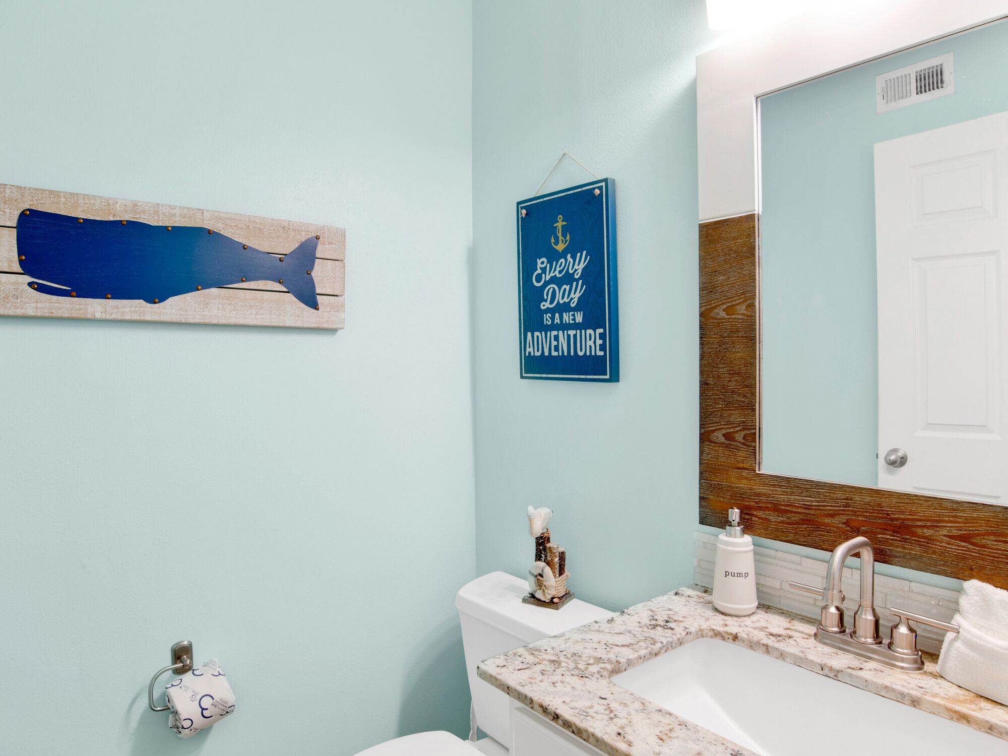 A bathroom with a light blue wall, a wooden shark artwork, a motivational quote, a marble sink, and a white toilet.