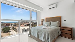 A modern bedroom with a large window, a bed with a wooden headboard, a nightstand, and a view of the cityscape. - MONYXBNB HOTEL (Puerto Vallarta)