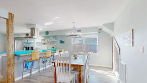 A modern kitchen with a dining area, featuring a large wooden table and white chairs, a blue countertop, and a view of the outside. - Wet Bar, Outdoor Games, Gulf Views, Pet Friendly (Port Aransas)