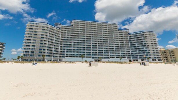 Un grande hotel a più piani con facciata curva, situato su una spiaggia sotto un cielo azzurro e nuvole sparse.