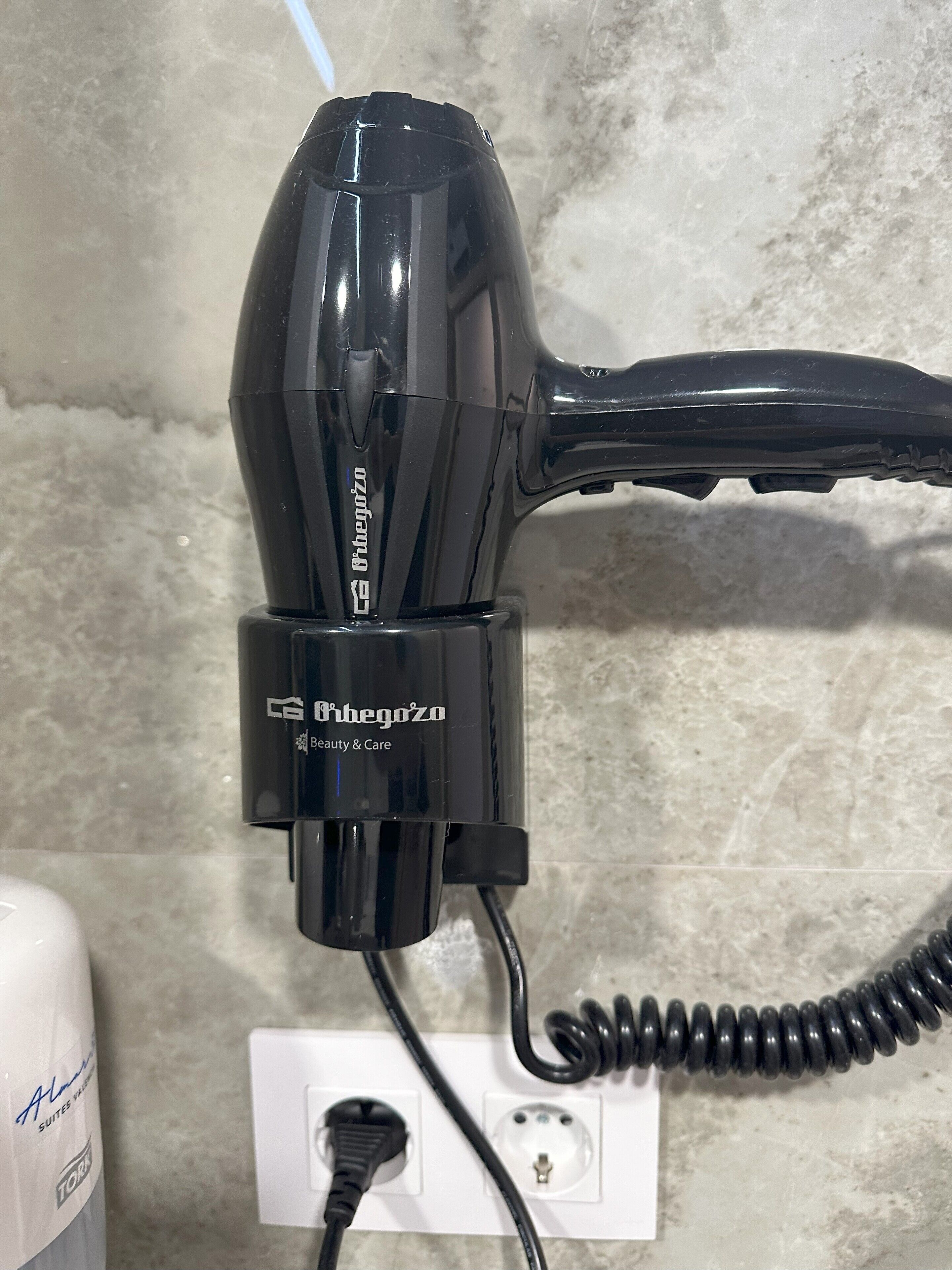A black hair dryer plugged into a white electrical outlet on a textured wall.