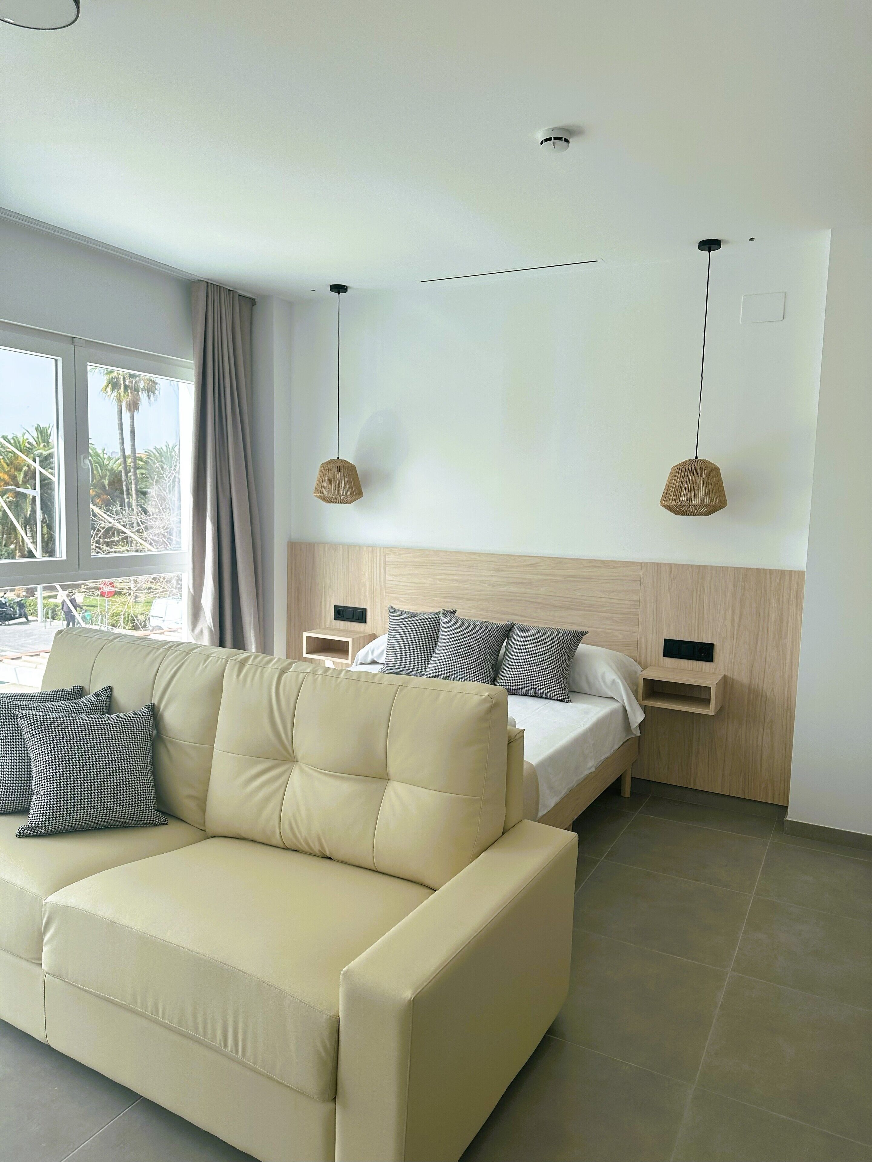 A modern hotel room with a beige sofa, a bed with grey and white bedding, two pendant lights, and a window with curtains.