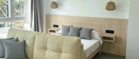 A modern hotel room with a beige sofa, a bed with grey and white bedding, two pendant lights, and a window with curtains.