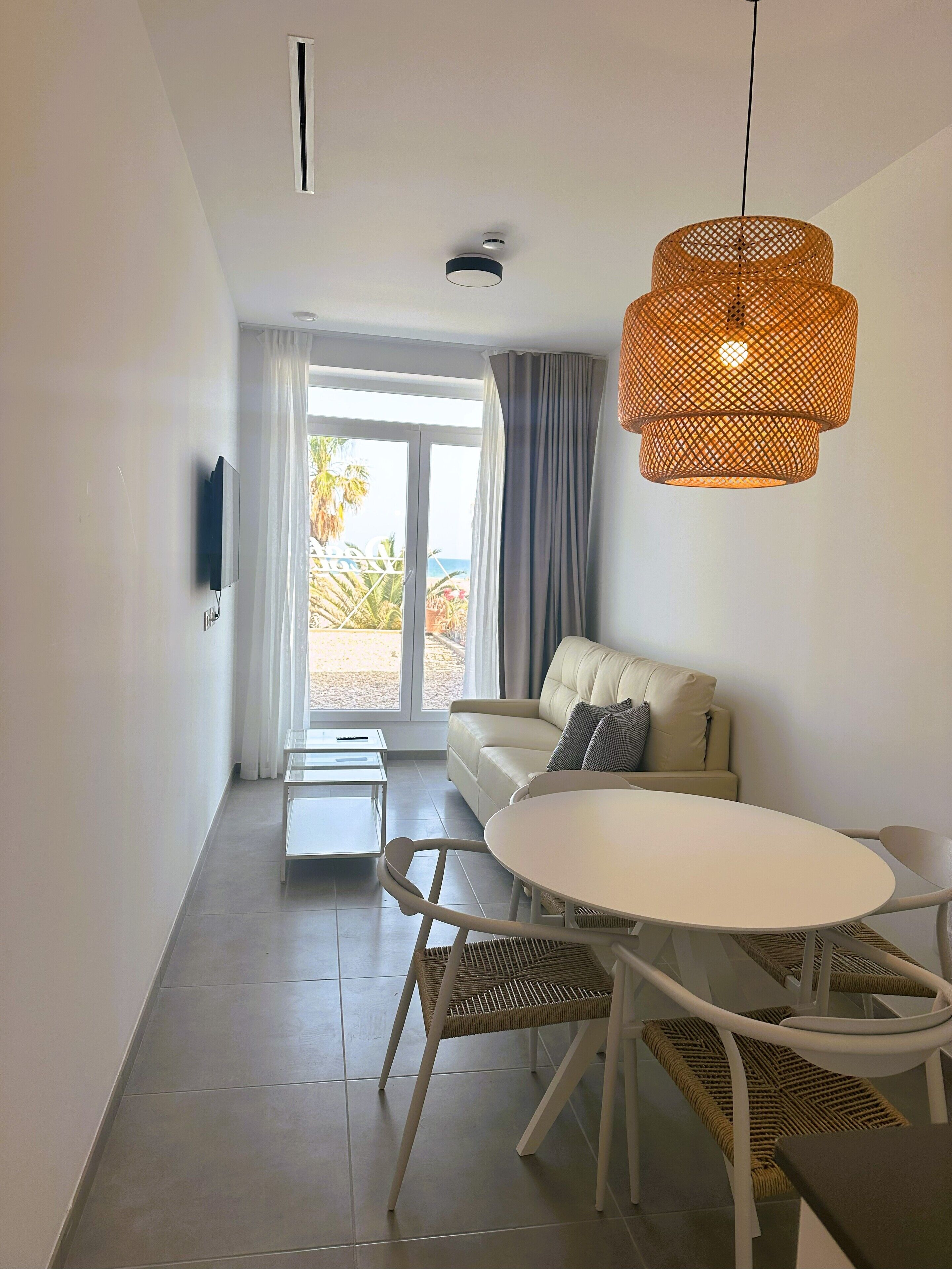 Una sala de estar moderna con un sofá, una mesa de comedor con sillas, un televisor montado en la pared y un ventanal con cortinas.