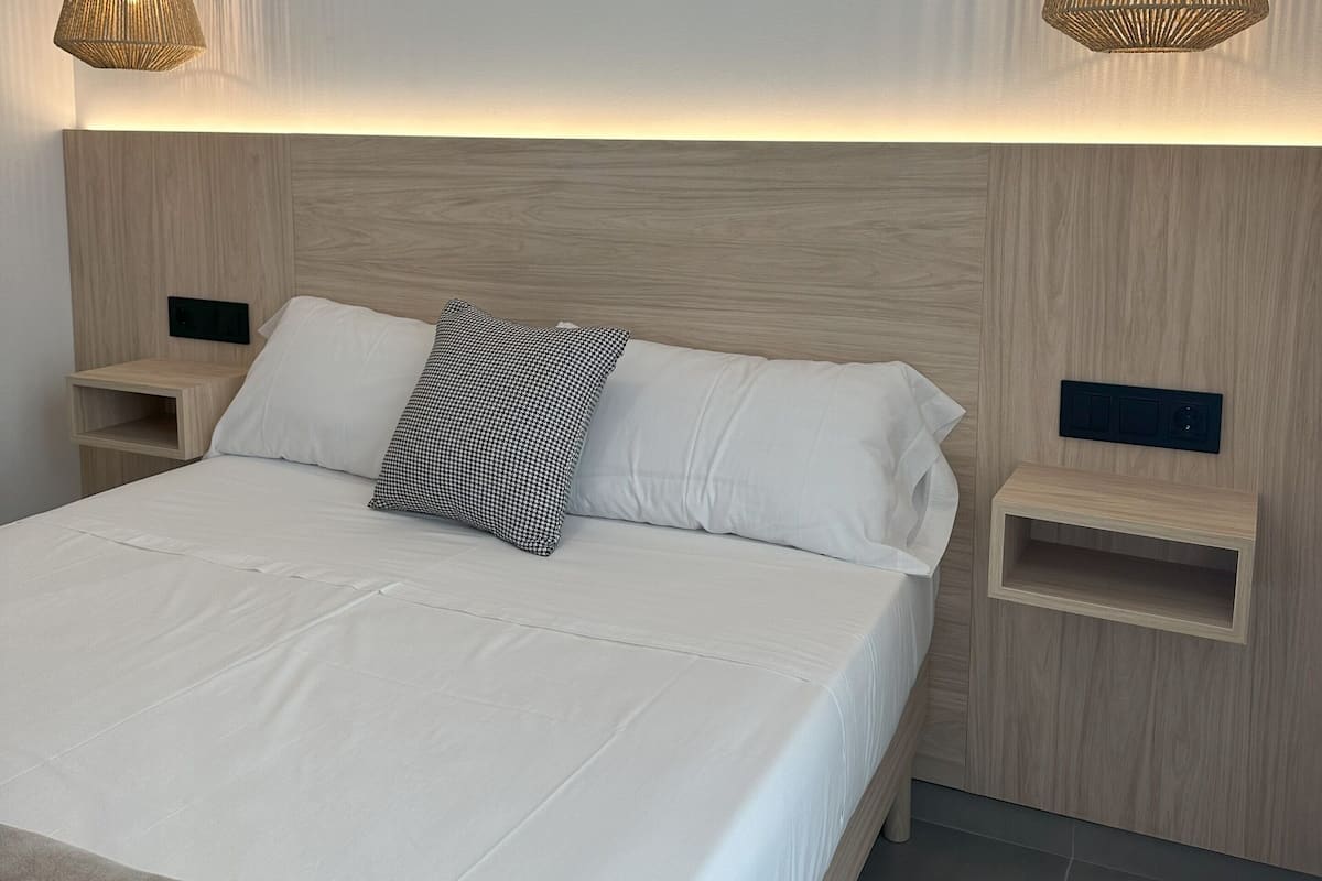 Une chambre à coucher moderne avec une tête de lit en bois, un grand lit avec une literie blanche, deux tables de chevet et deux lampes suspendues.