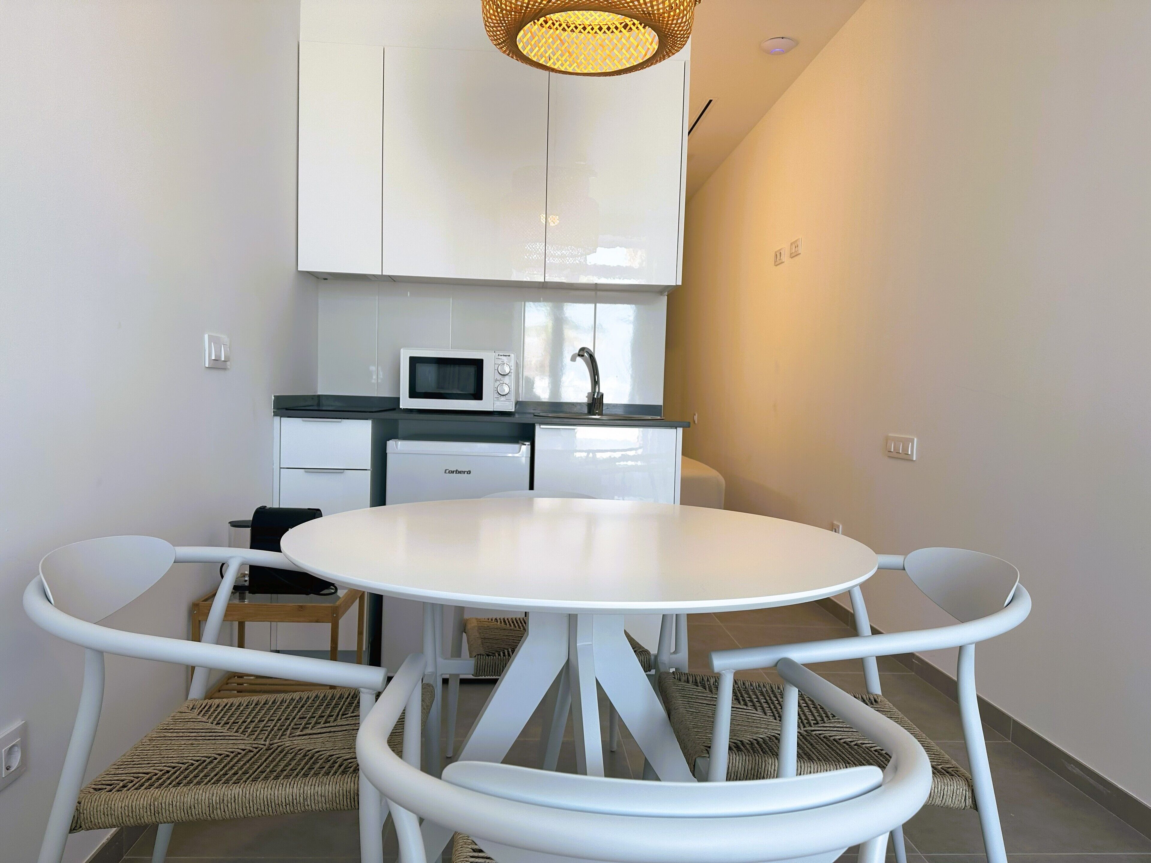 Une cuisine compacte avec des meubles blancs, un four à micro-ondes et un évier. On y trouve une table ronde blanche avec quatre chaises, et une chaise en osier est placée à côté de la table.